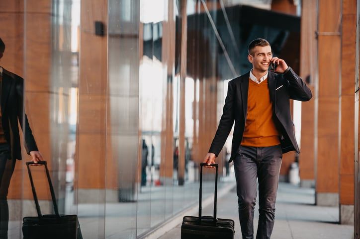 A man in business casual clothing walks with a rolling suitcase while talking on the phone.