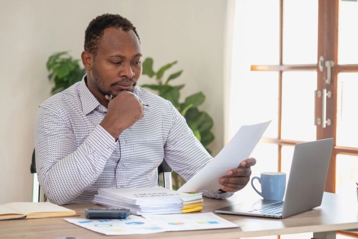 A man sits at a desk with a laptop while looking at a pile of business documents.