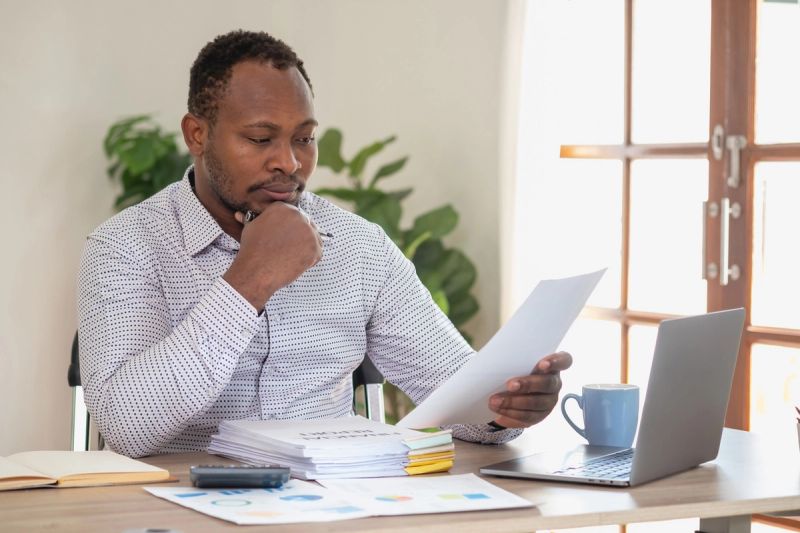 A man sits at a desk with a laptop while looking at a pile of business documents.