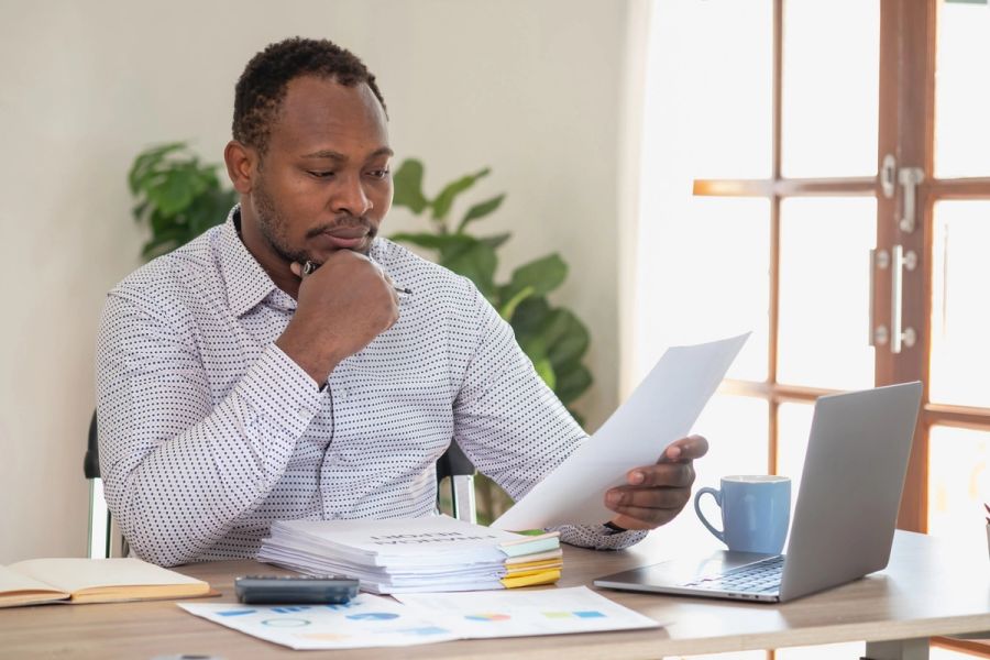 A man sits at a desk with a laptop while looking at a pile of business documents.