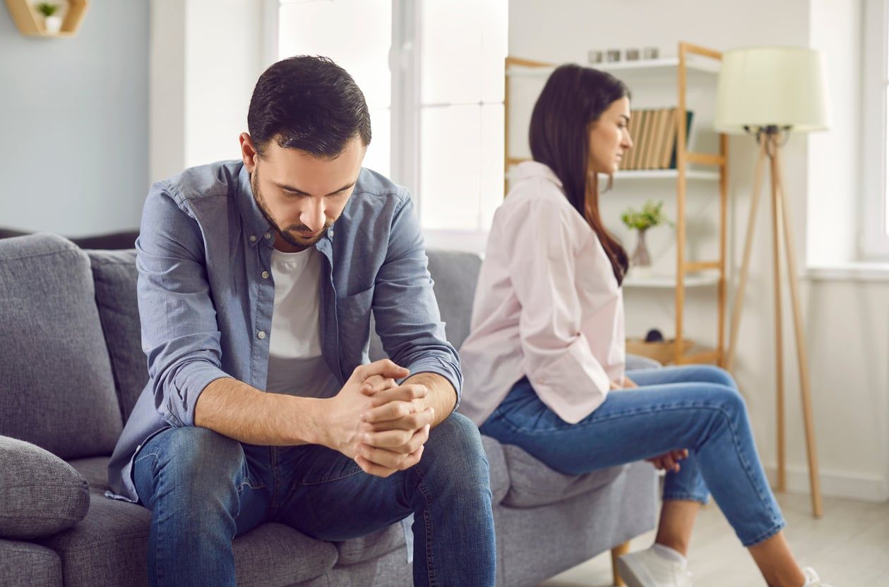 An upset married man and woman sit on a sofa looking away from each other.