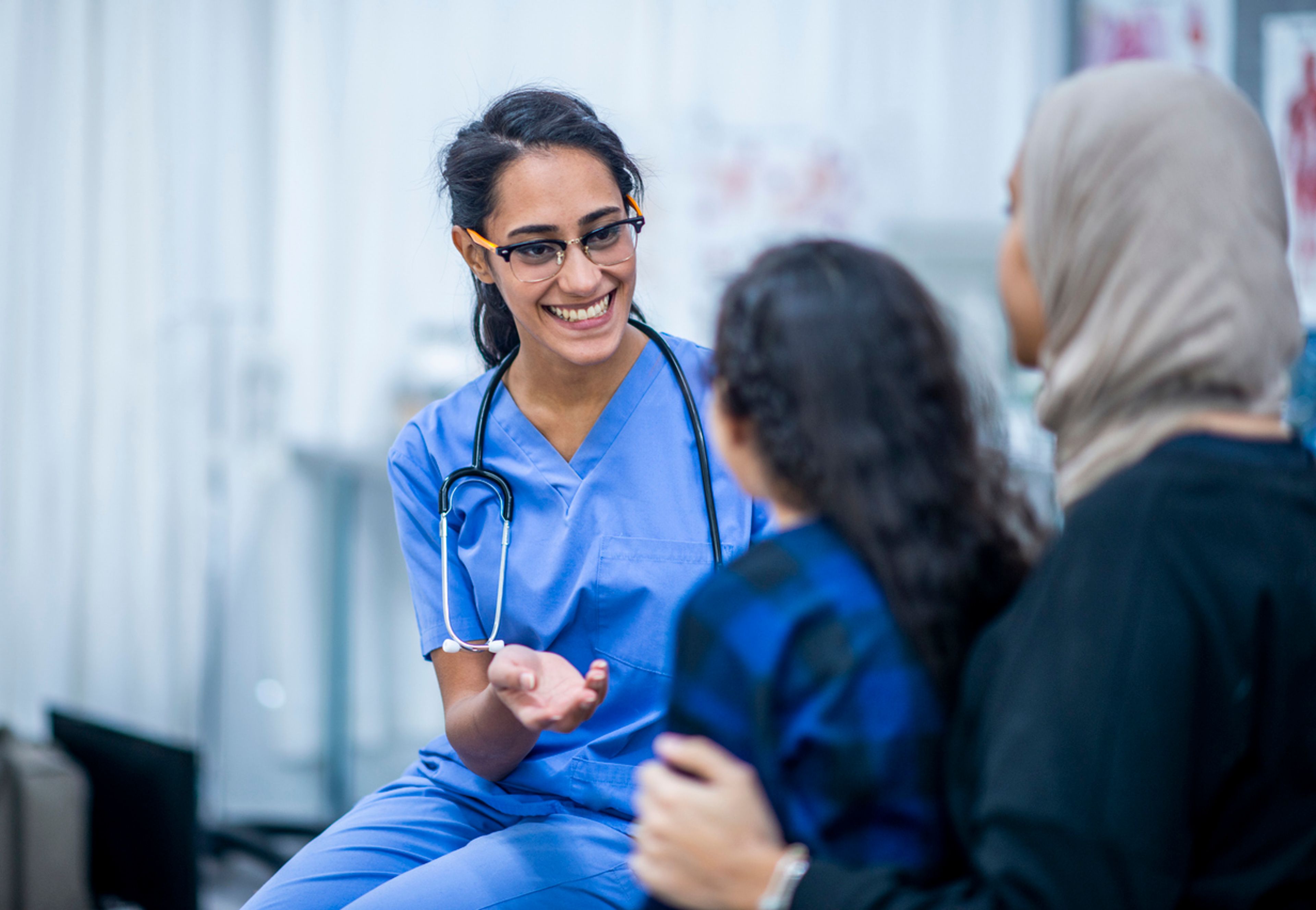 A seated, female registered nurse in scrubs talks with a child.