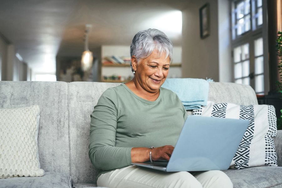 An older woman sits on a couch and smiles while looking at her laptop.