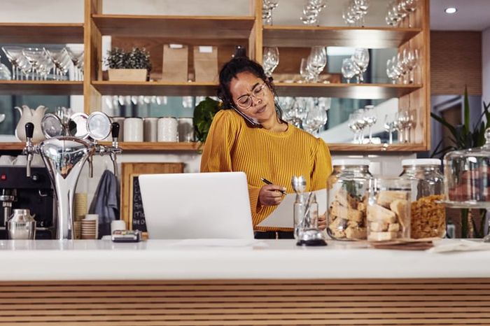 A restaurant owner woman talks on the phone while looking at a laptop.