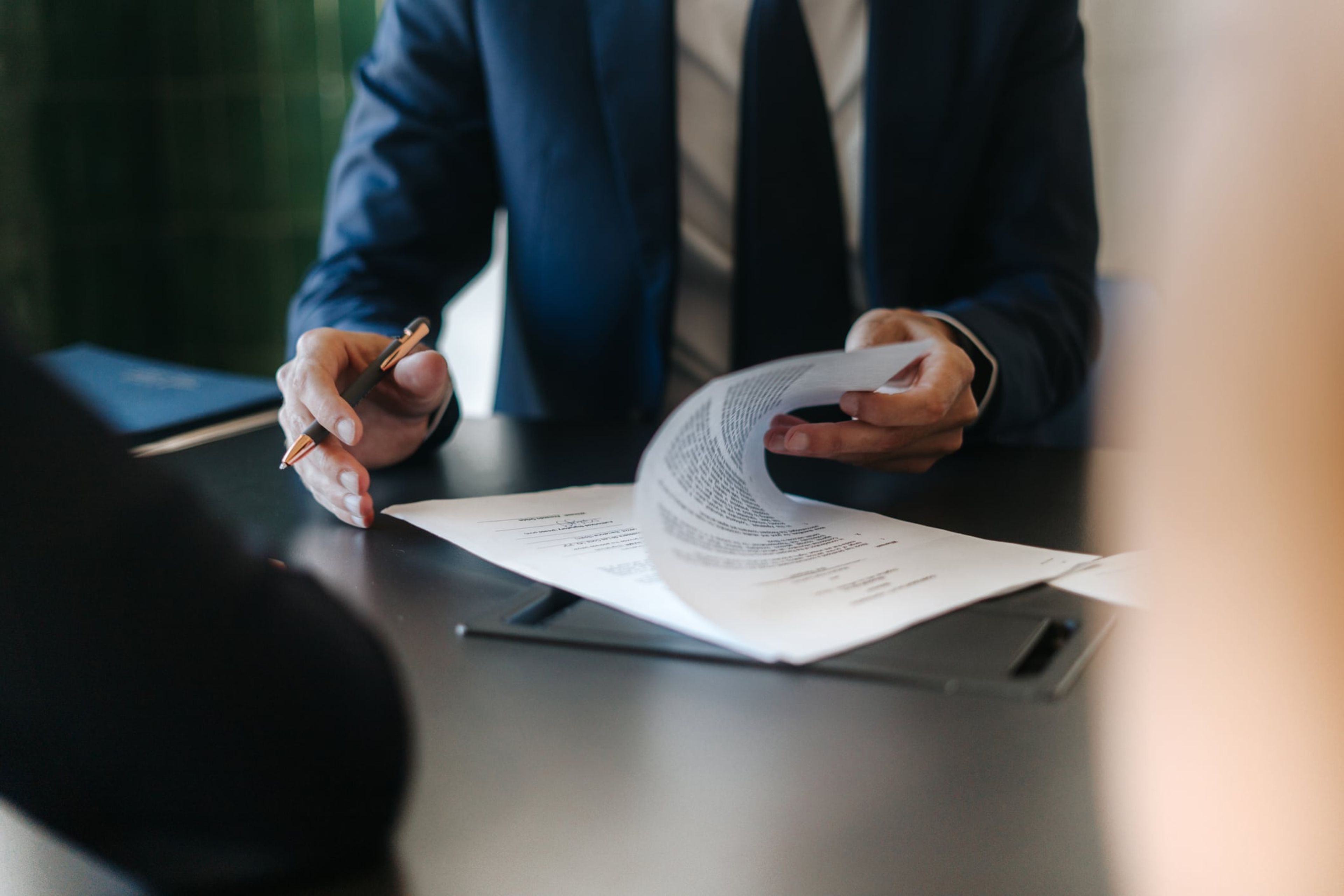 A man in a blue suit sits at a desk with a pen in one hand, while looking over a contract.