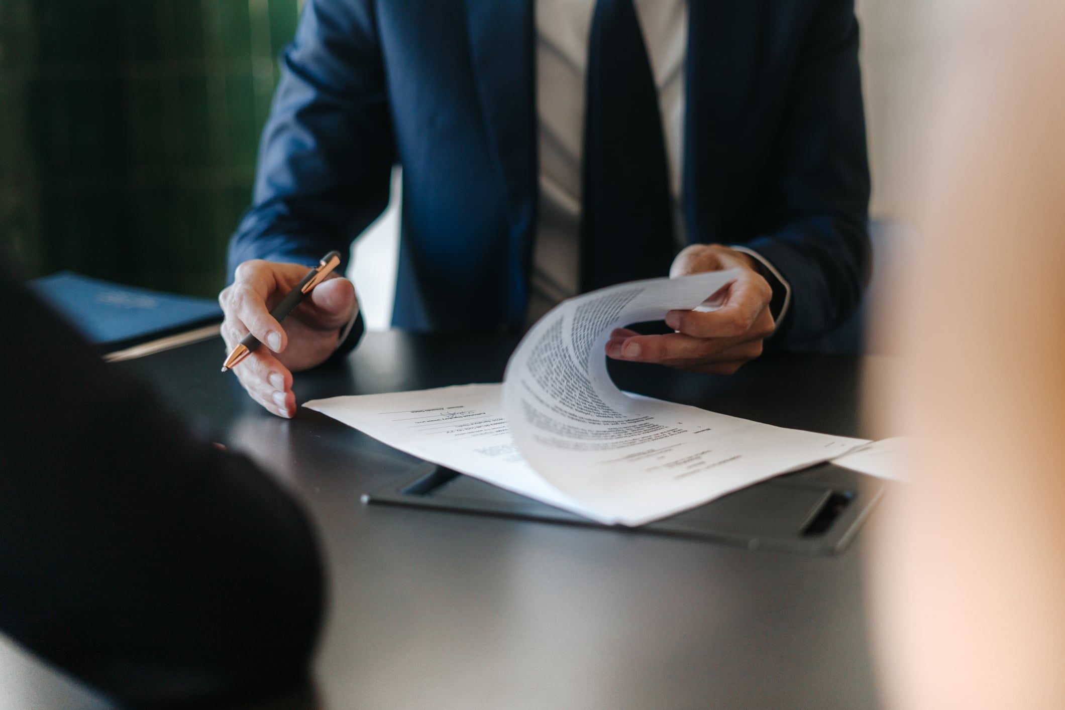 A man in a blue suit sits at a desk with a pen in one hand, while looking over a contract.