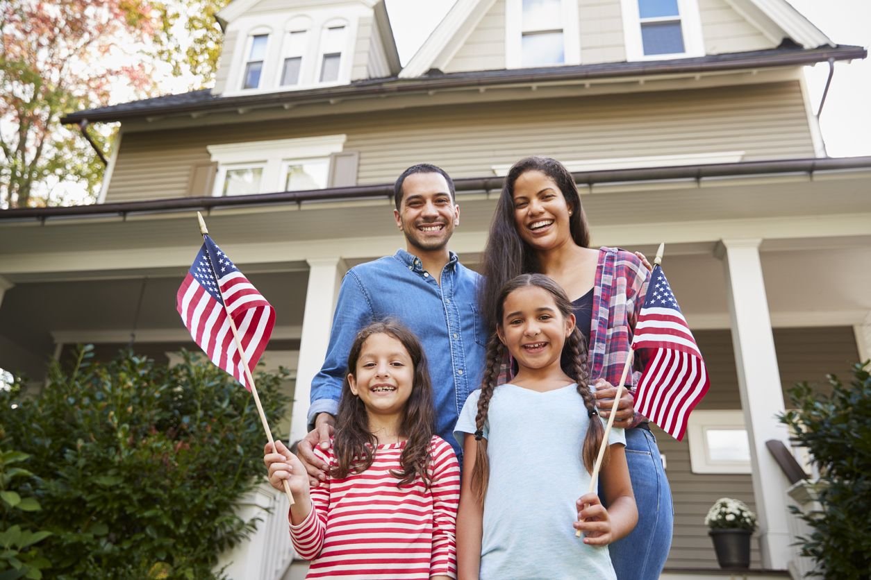 Family of four standing in front of house with both daughters holding an American flag.