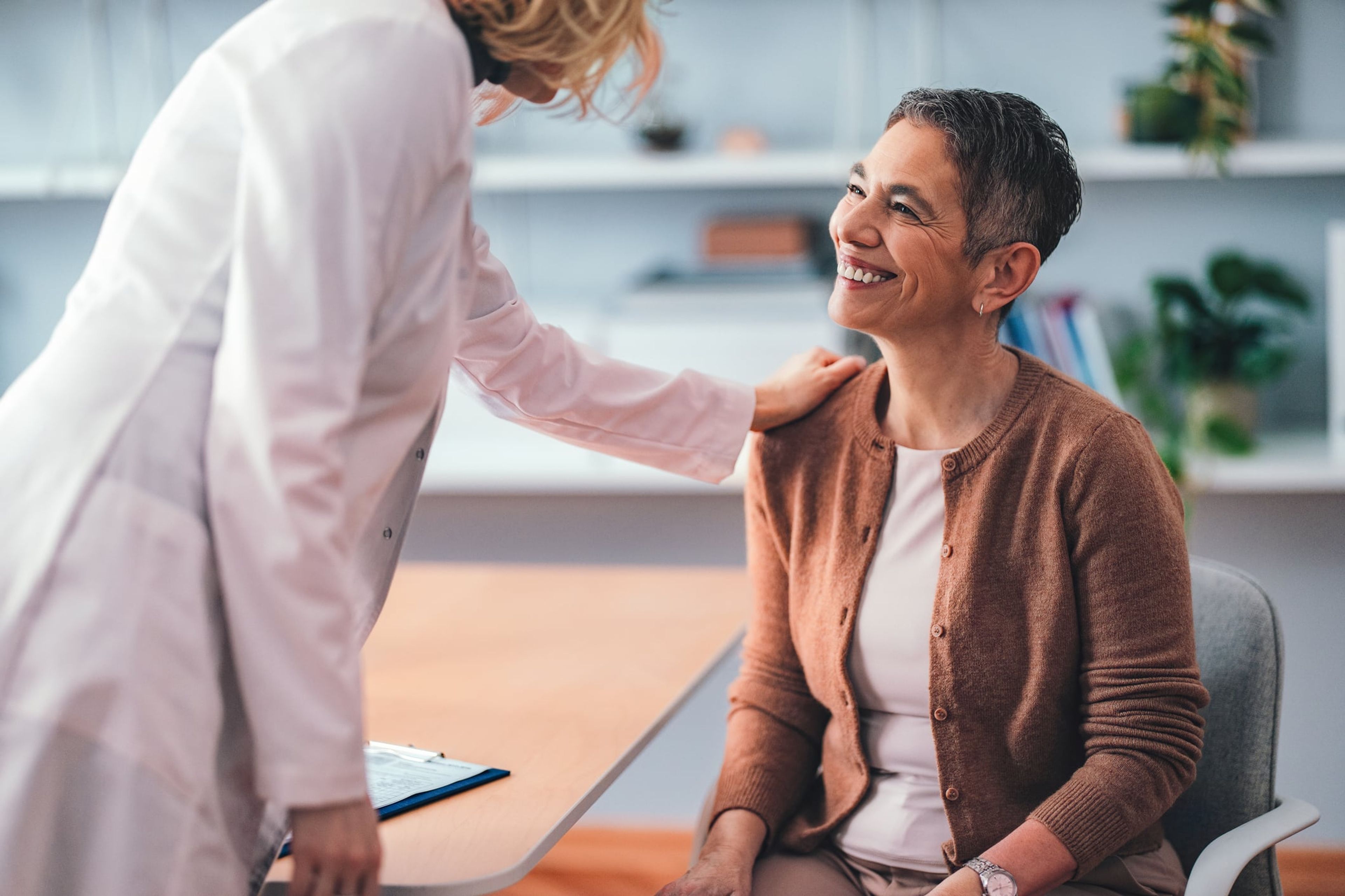 A woman sits in a front of a physician for a Green Card medical exam, as the doctor puts a hand on her shoulder.