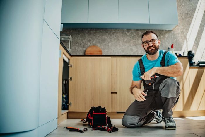 A male plumber kneels in a kitchen with tools in front of him.