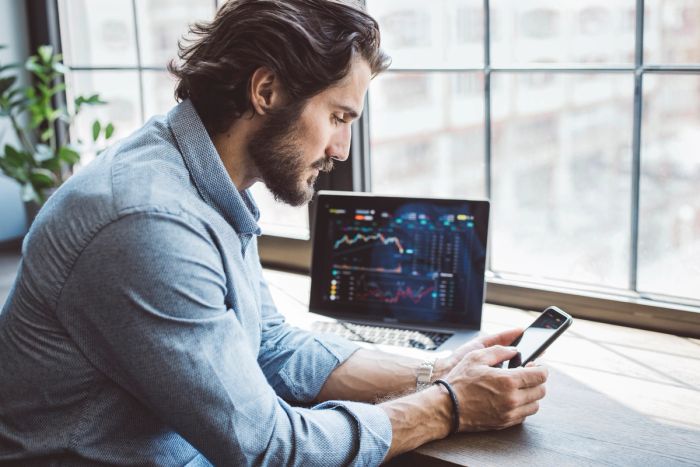 A male financial investor sits and looks at his phone while a laptop next to him shows stock projections.