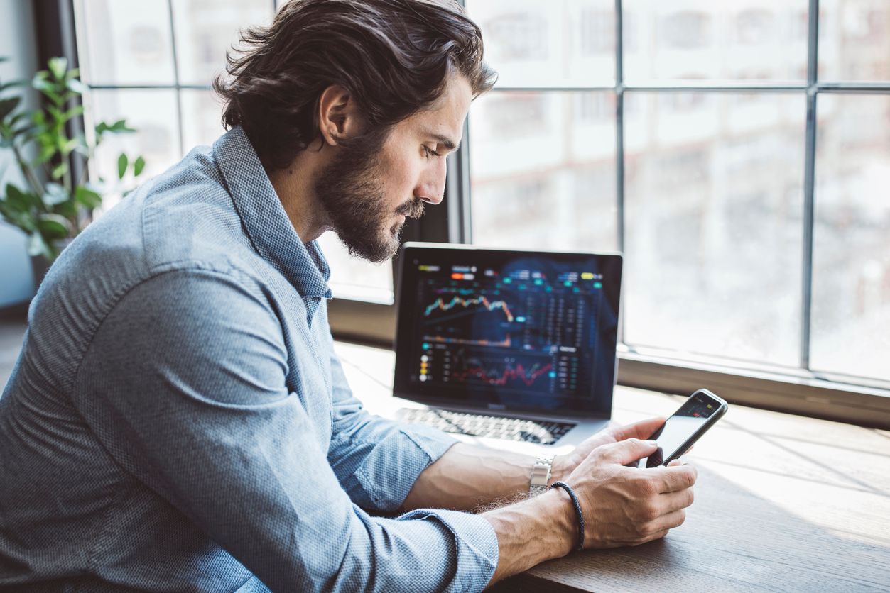 A male financial investor sits and looks at his phone while a laptop next to him shows stock projections.