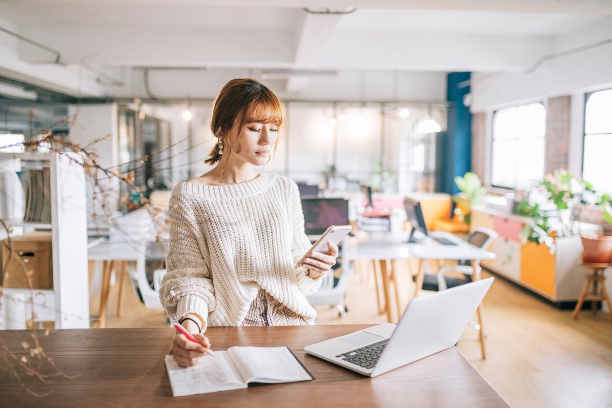 A woman stands at a table and writes in a notebook while looking at her phone.