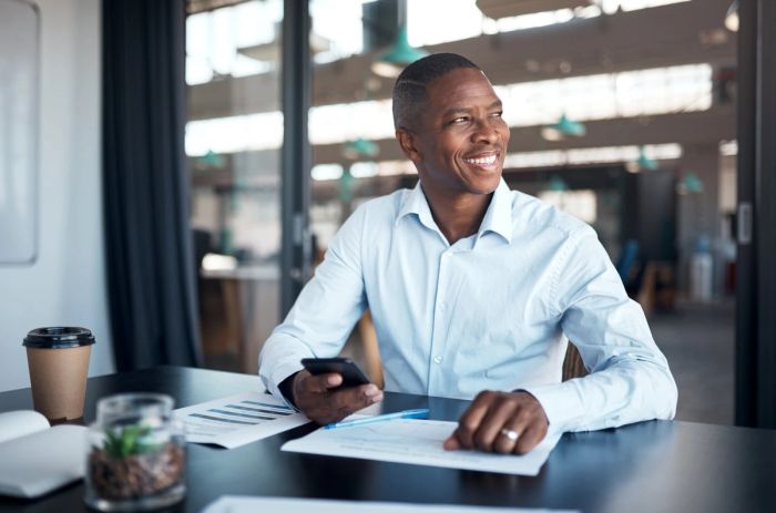 An older businessman smiles while sitting at a desk in front of some papers and holding his smartphone.