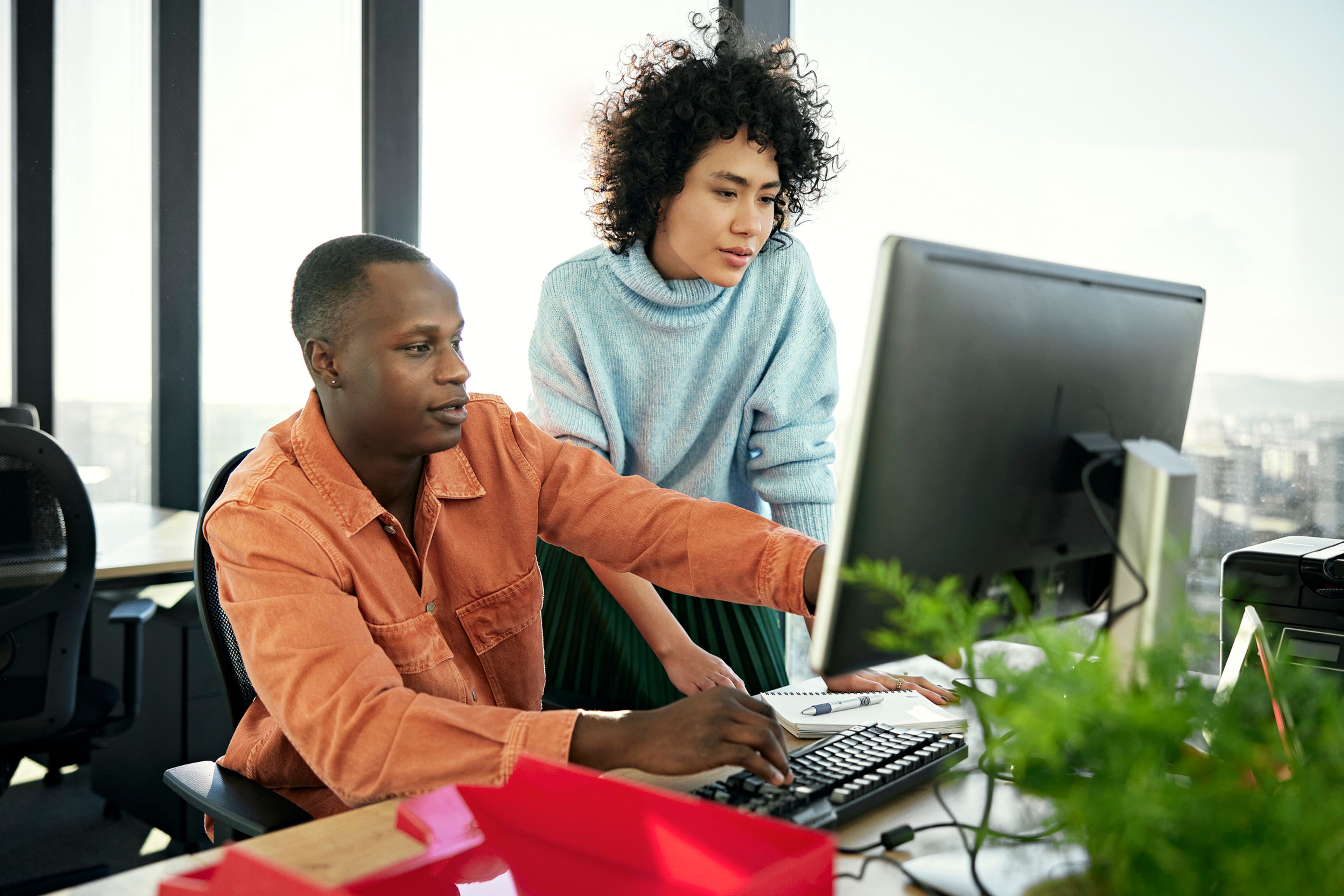 Waist-up view of casually dressed associates collaborating as they sit and stand at flexible workstation, looking at desktop PC monitor.