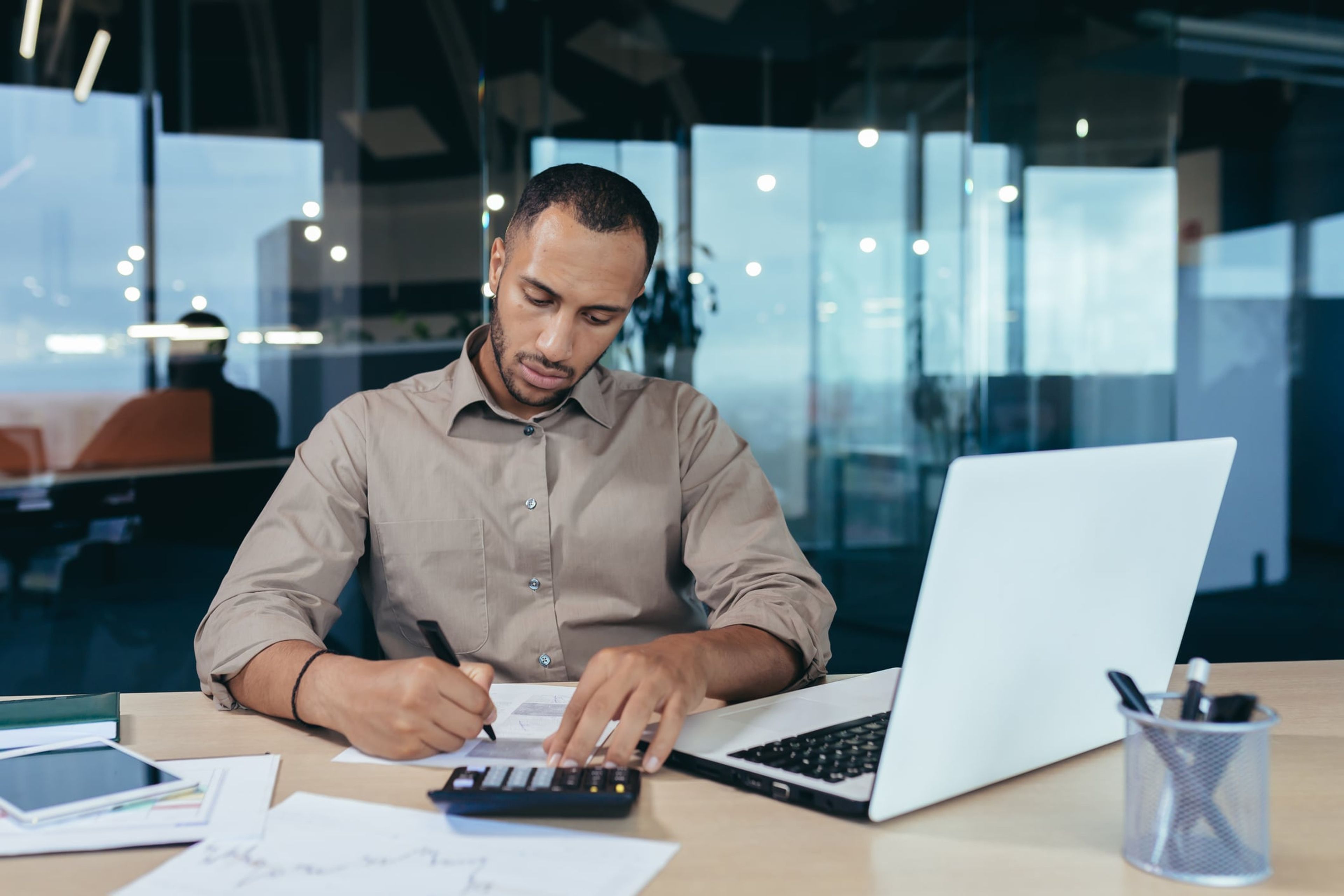 A young international businessman sits at a desk and handwrites notes with a laptop and calculator in front of him