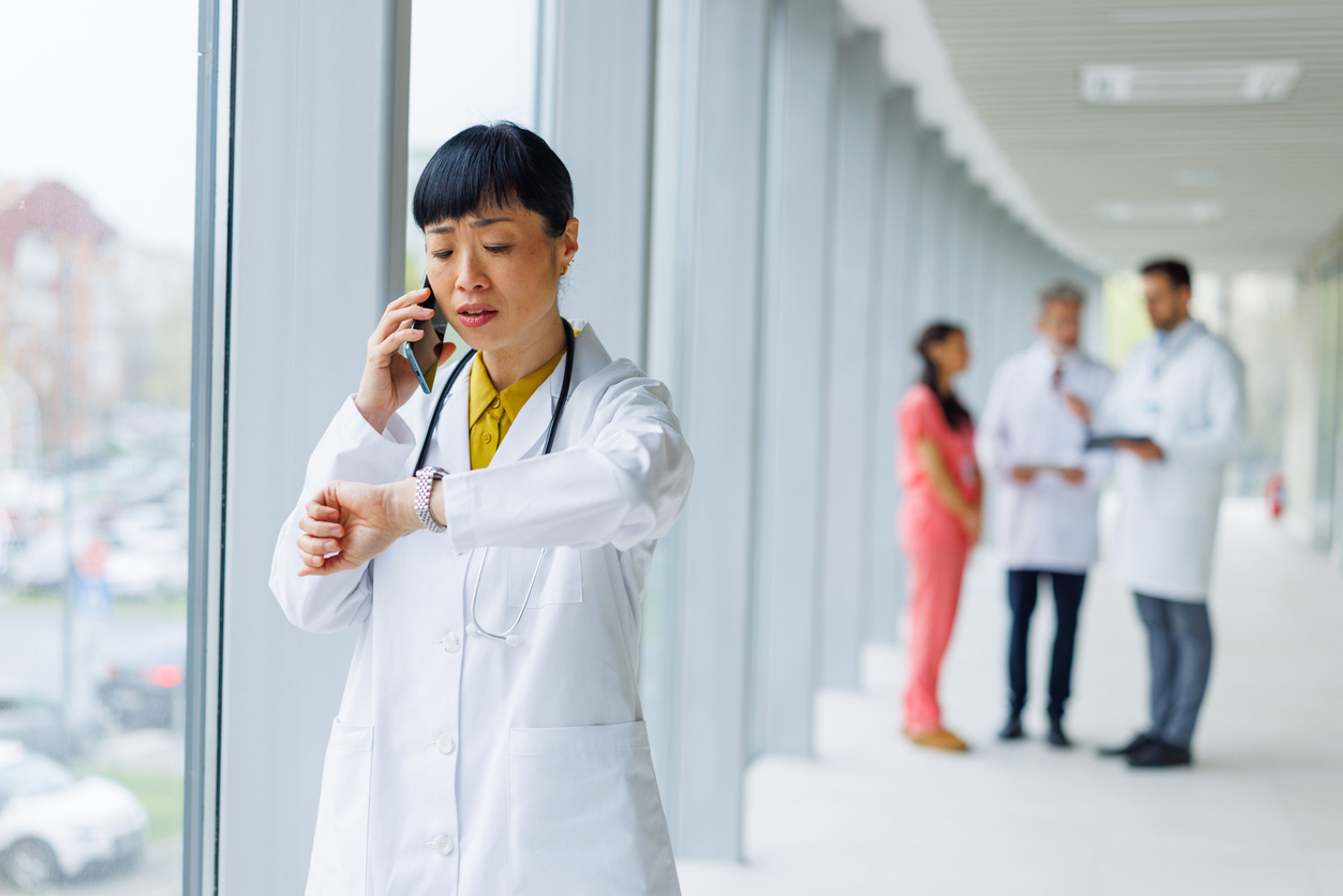 A female doctor in a white lab coat is talking on phone while checking her watch.