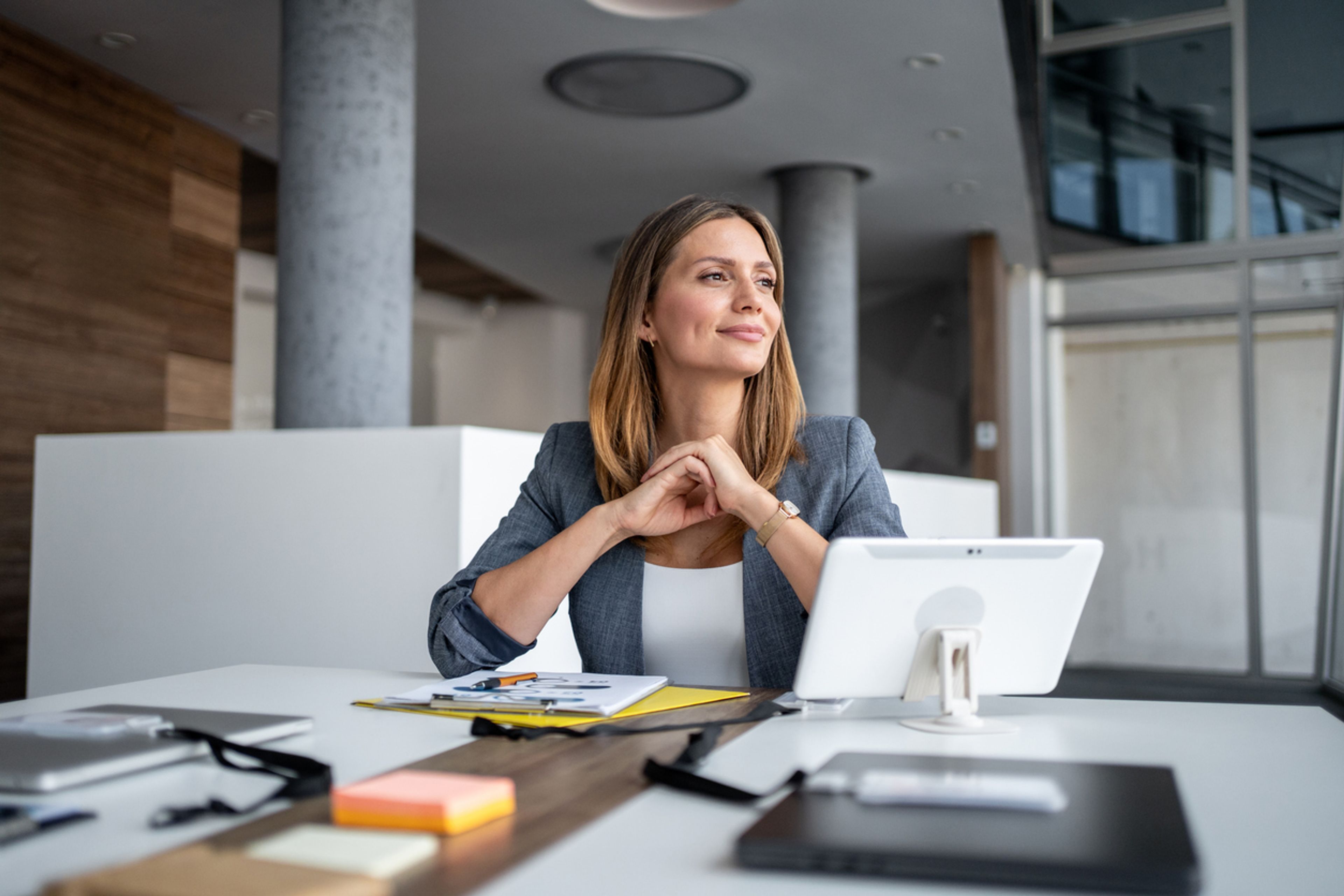 Businesswoman sitting at a modern office desk, smiling and gazing thoughtfully into the distance.