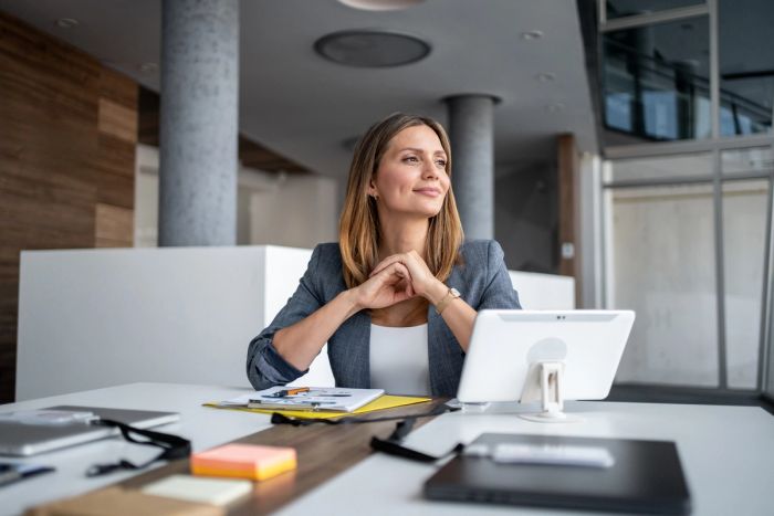 Businesswoman sitting at a modern office desk, smiling and gazing thoughtfully into the distance.