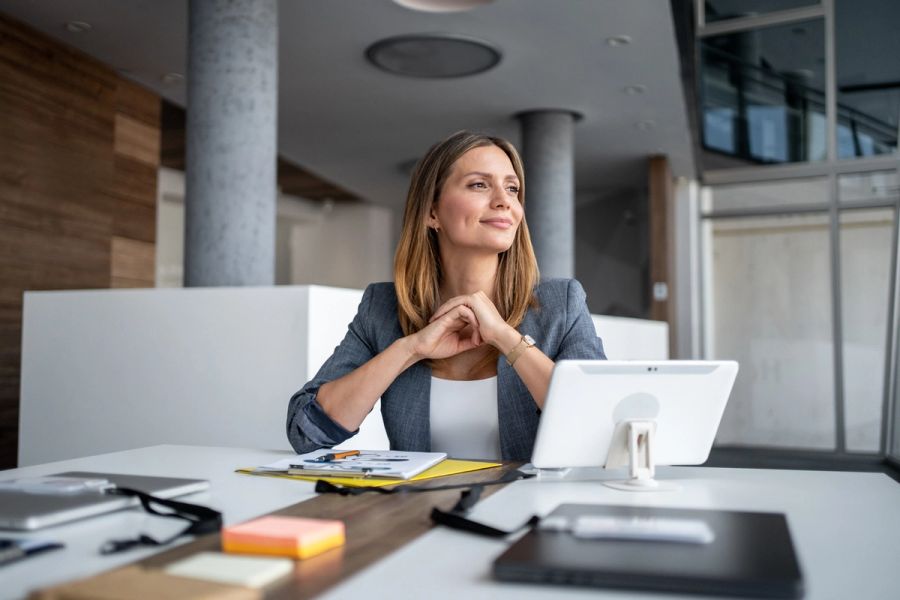 Businesswoman sitting at a modern office desk, smiling and gazing thoughtfully into the distance.