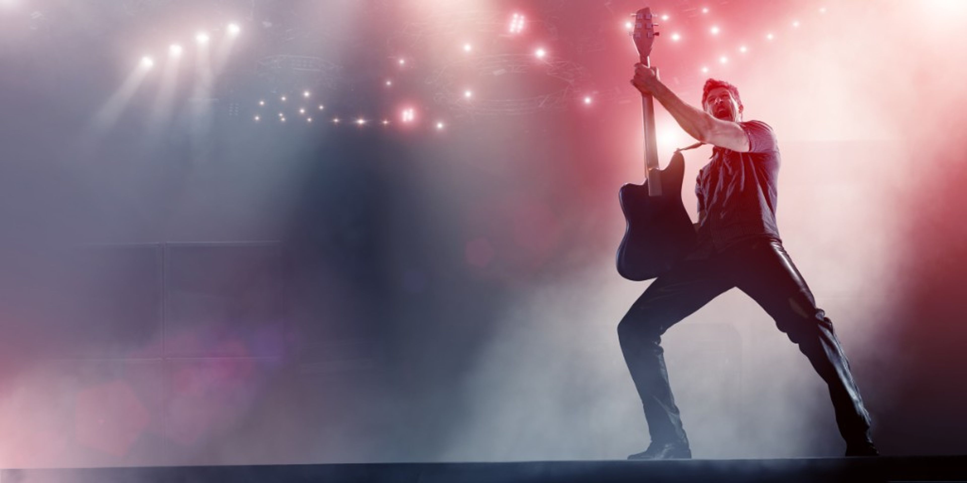 A performer on a concert stage holding an electric guitar amid dramatic stage lighting and haze.