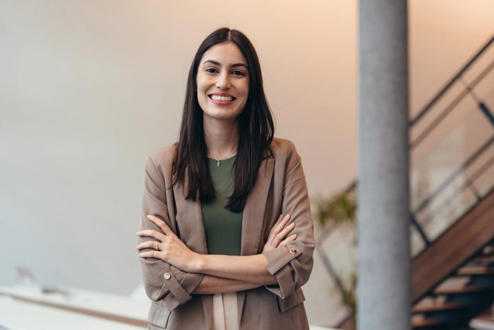 Confident young businesswoman smiling at the camera while standing with arms crossed in a sleek modern office.