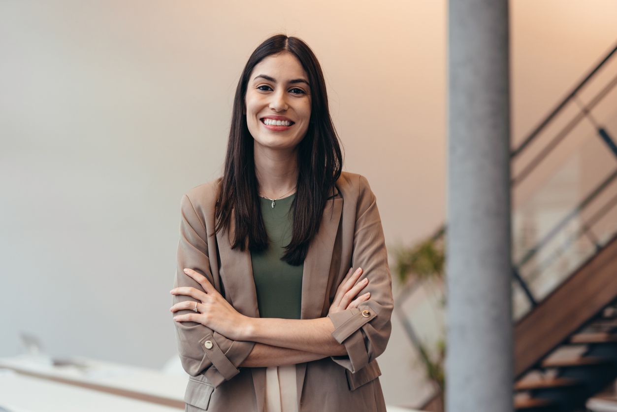 Confident young businesswoman smiling at the camera while standing with arms crossed in a sleek modern office.