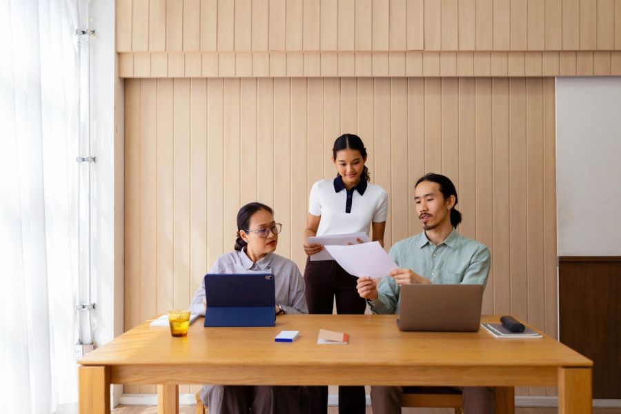 Three colleagues collaborate over laptops and documents at a wooden table in a bright modern workspace.