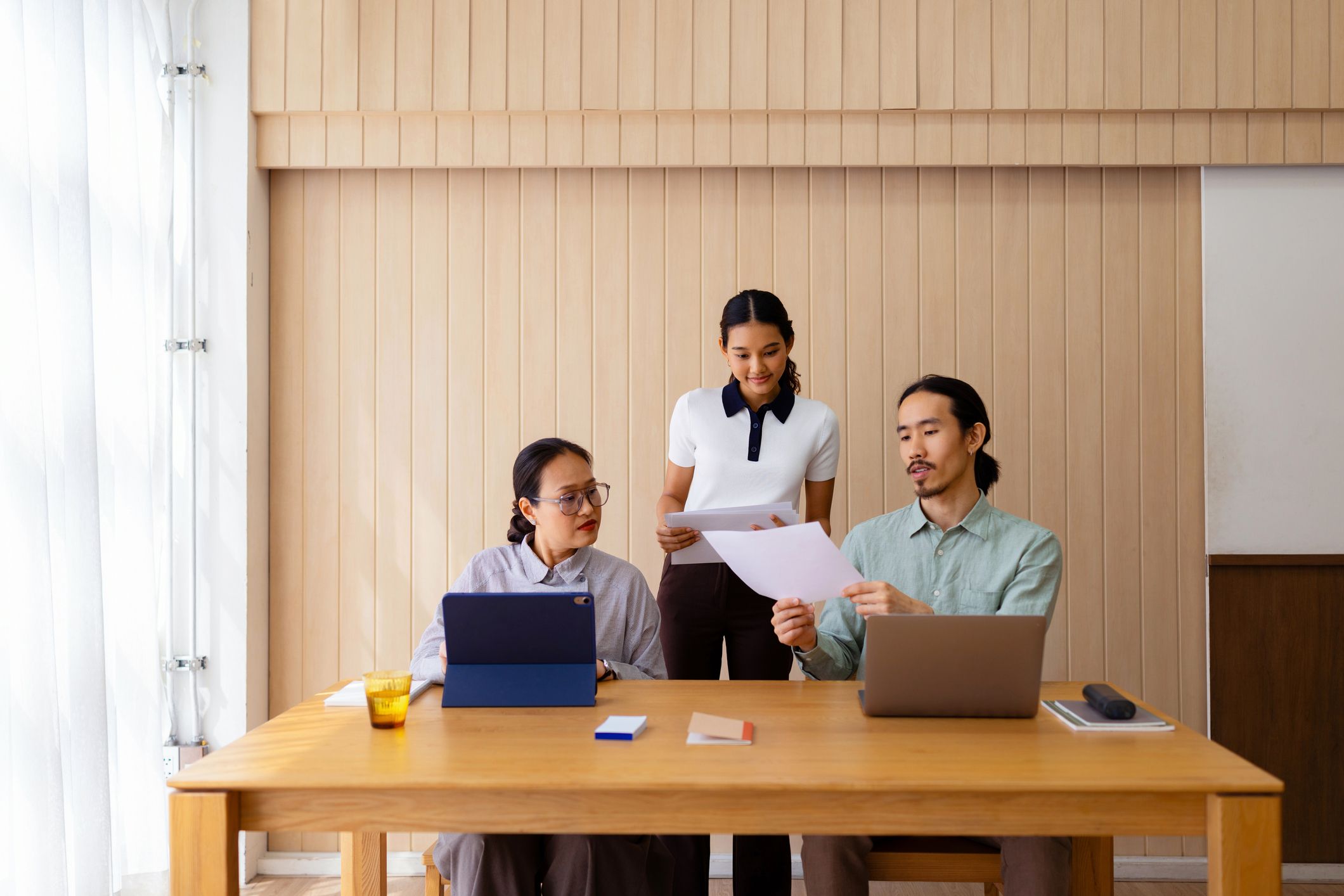 Three colleagues collaborate over laptops and documents at a wooden table in a bright modern workspace.
