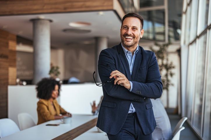A successful businessman stands in an office smiling.