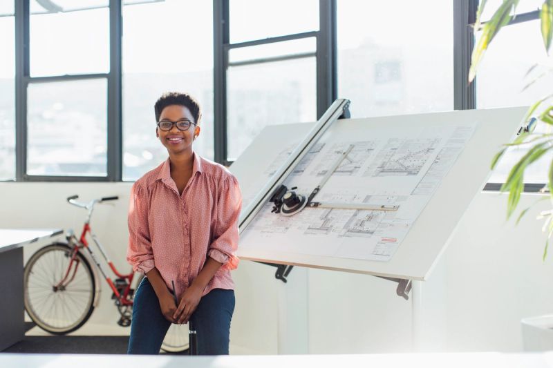 A young woman sits in front of large papers with architectural designs.