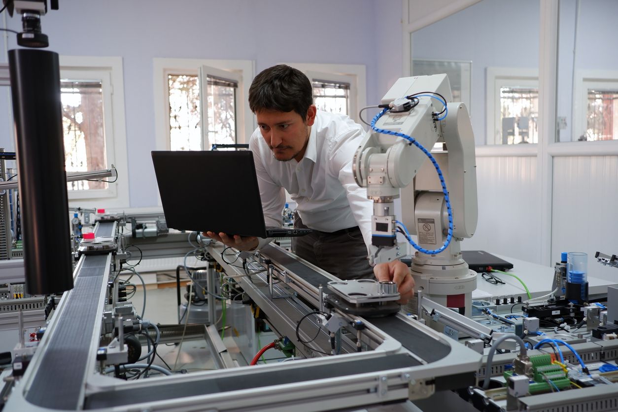 A man looks at a laptop while leaning over a table with a robotic arm and other electronic equipment.