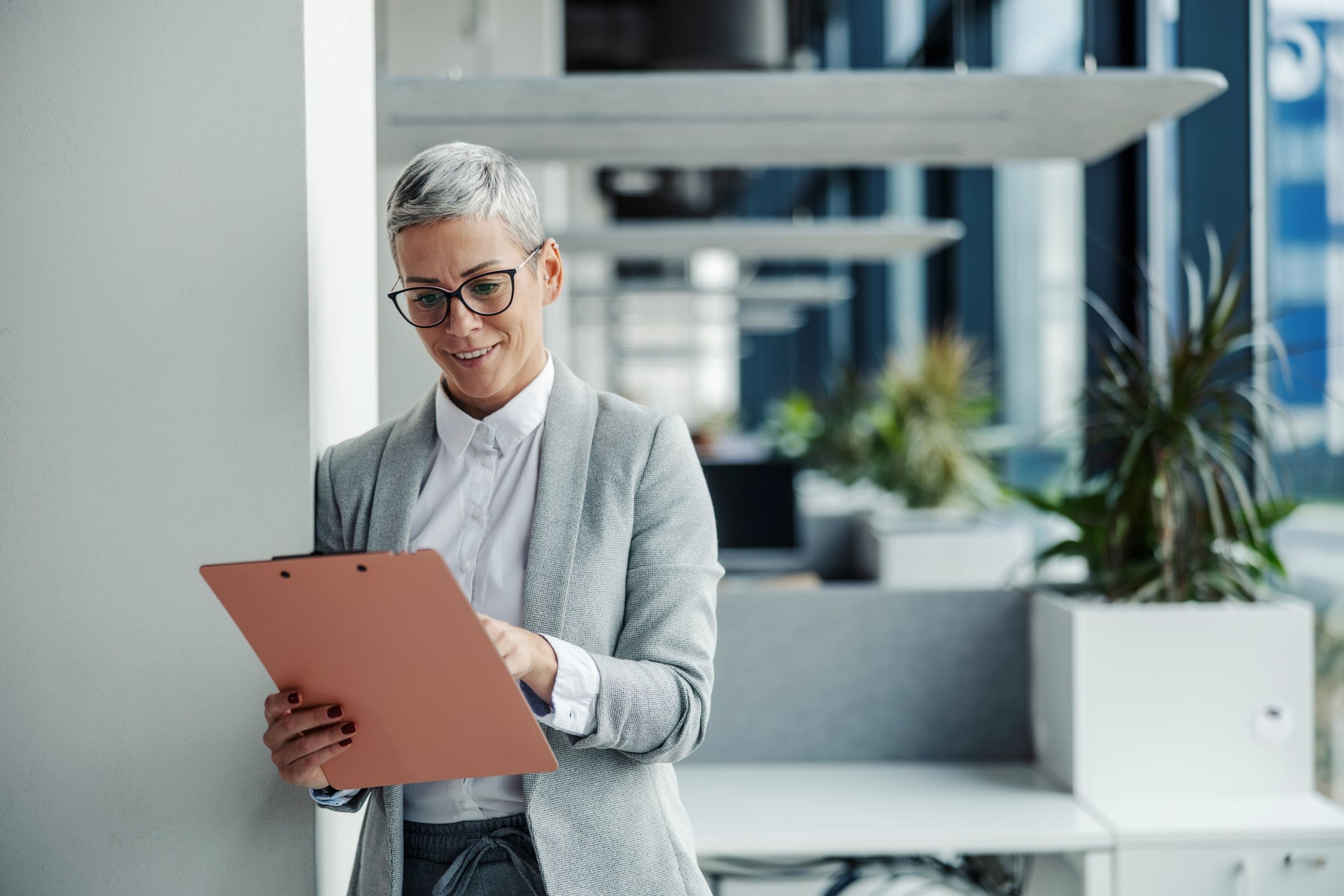 A businesswoman stands in an office smiling as she reviews documents on a clipboard.