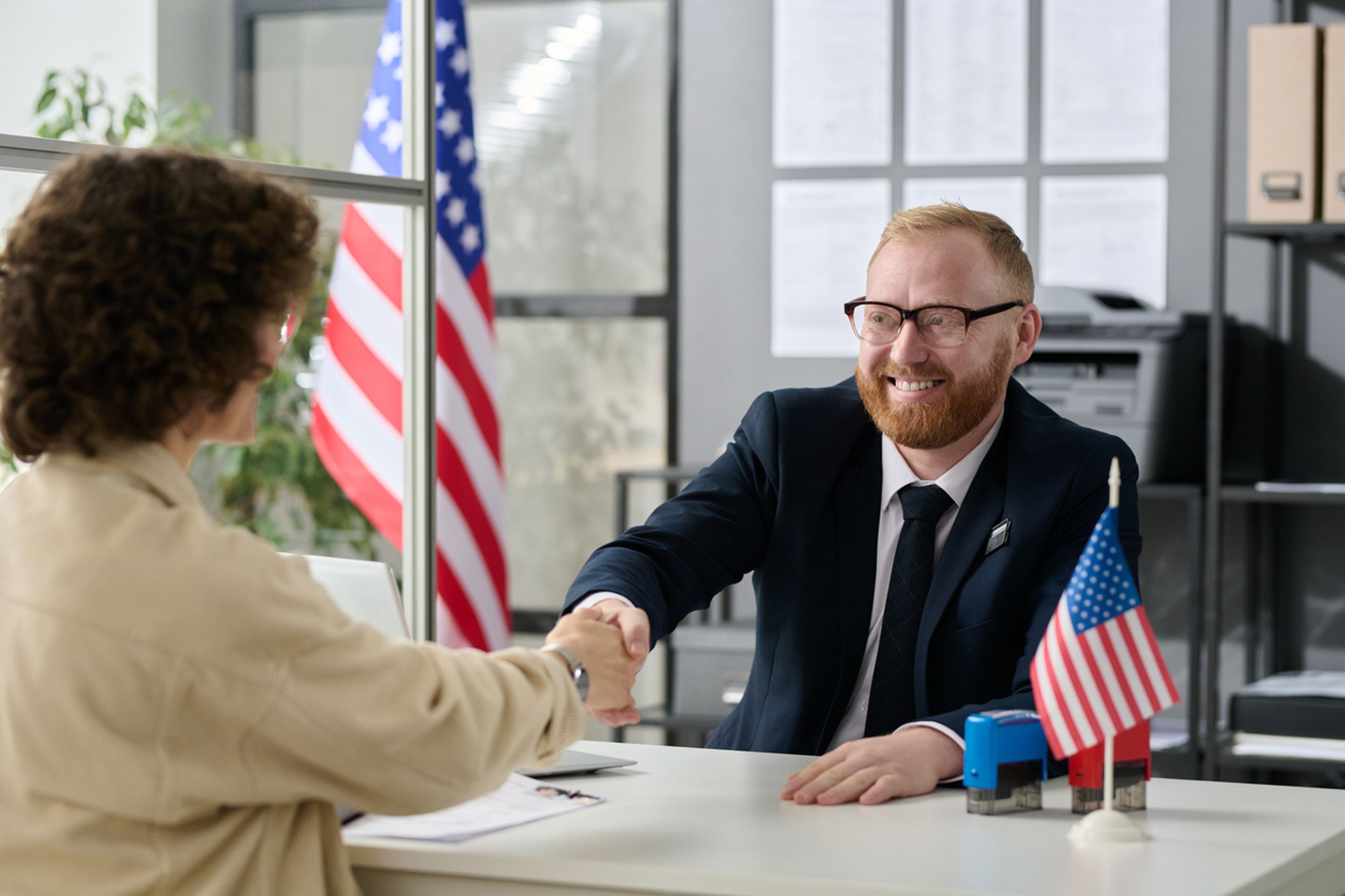 A male, U.S. immigration officer in a suit shakes hands over a desk with a woman over.