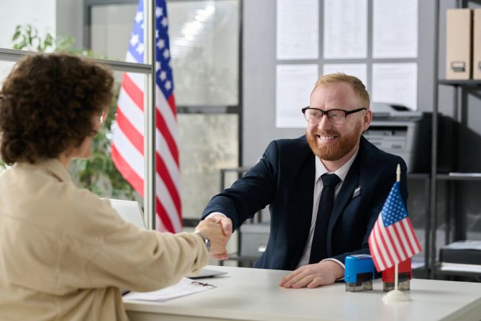 A male, U.S. immigration officer in a suit shakes hands over a desk with a woman over.
