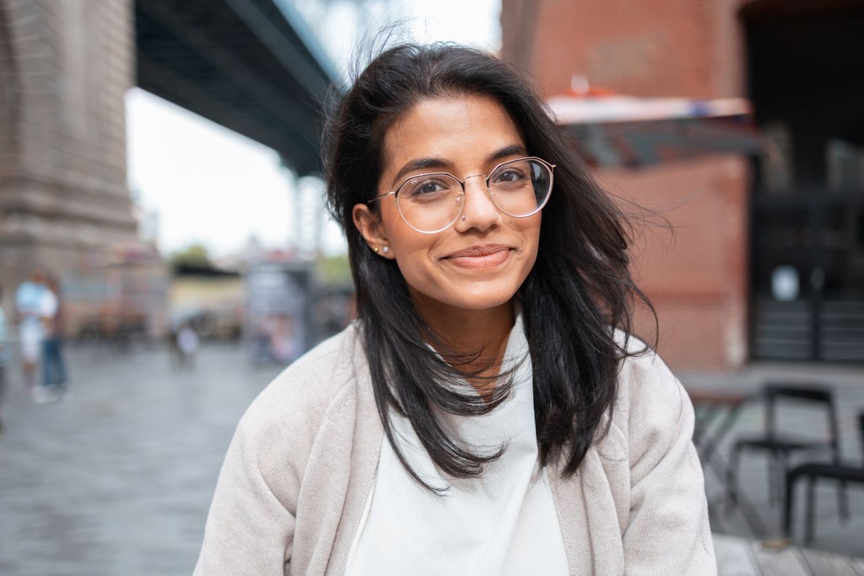 Young adult woman wearing glasses and smiling, standing outdoors with the Brooklyn Bridge in the background.