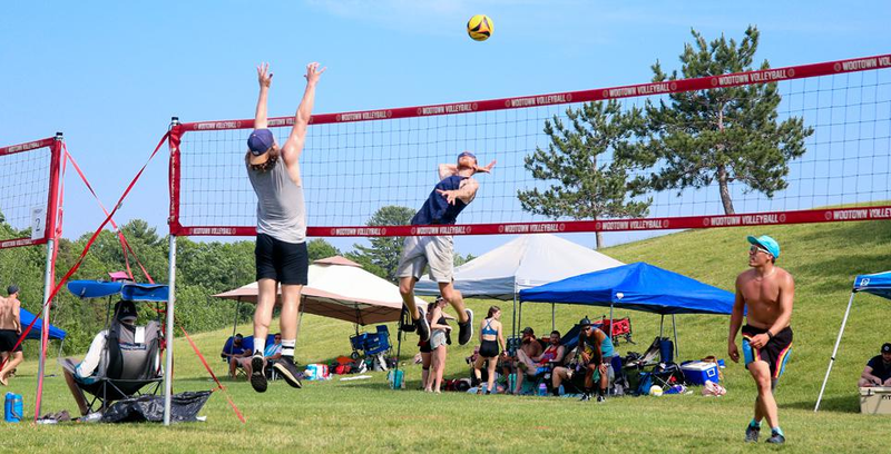 Two men are jumping near a volleyball net during an outdoor game on a sunny day. Tents and spectators are visible in the background on a grassy field.