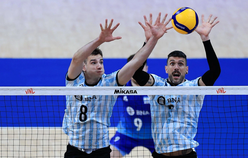Two volleyball players in blue and white jerseys are jumping to block a ball above the net during a match.