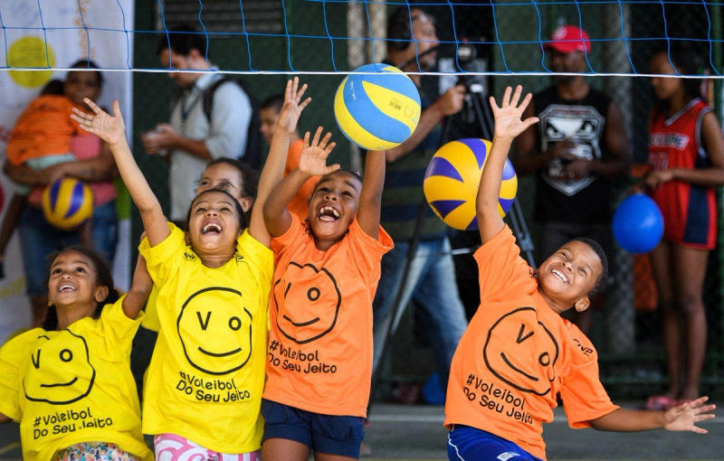 Children in bright yellow and orange shirts joyfully reach for volleyballs above a net, with onlookers and balloons in the background.