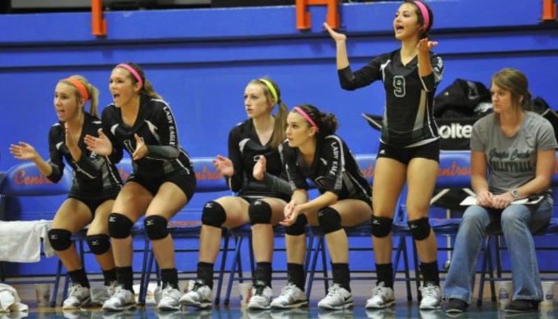 A group of five volleyball players in black uniforms sits on the bench, actively reacting to the game with clapping and gestures. Another person, in casual attire, calmly holds a clipboard beside them.