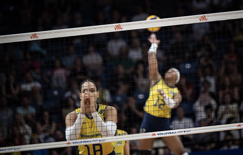 Two volleyball players in yellow jerseys are on a court. One is preparing to block near the net, while the other is jumping for a spike in the background. The scene is captured during a match with a crowd watching.