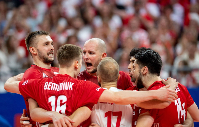 A group of volleyball players in red jerseys huddle together in a celebratory and intense moment on the court, with an excited crowd in the background.