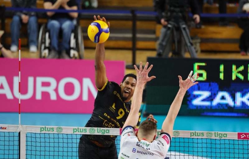 A volleyball player in a black jersey spikes the ball over the net during a match, while an opposing player in a white jersey attempts to block. Spectators and a cameraman are seen in the background.