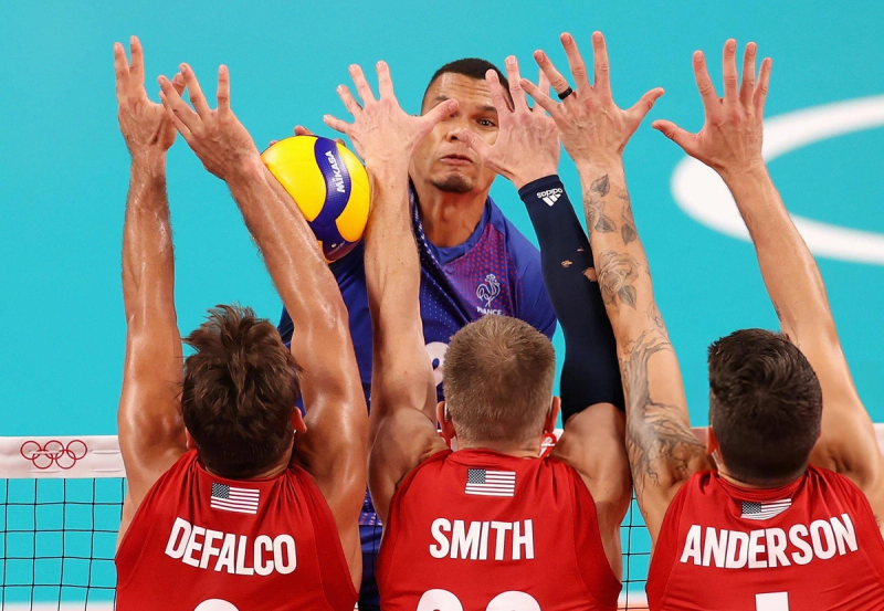 Volleyball game scene with a player in a blue jersey attempting a spike against three players in red jerseys attempting to block at the net.