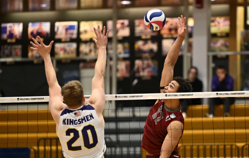 Two volleyball players at the net during a championship match. One player, in a light jersey with "Kingsmen 20" on the back, is attempting to block a spike from the opposing player in a dark jersey. The ball is visible above the net.