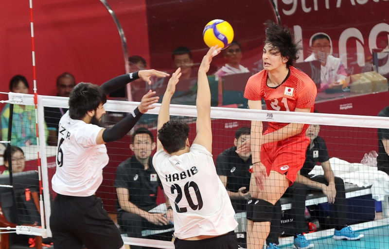 A volleyball player in a red jersey jumps to spike the ball over the net, while two opposing players in white jerseys attempt to block. Spectators and team staff are visible in the background.