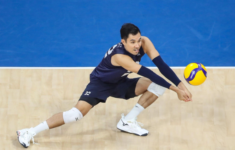 A volleyball player in a navy uniform performing a low dig on a court, poised with arms extended and knees bent, as a yellow and blue volleyball approaches.