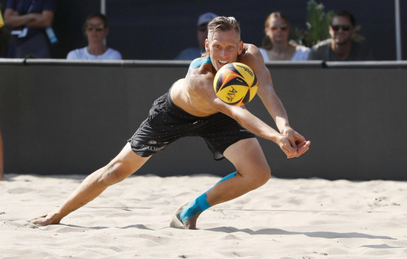 A male beach volleyball player dives in the sand to bump a volleyball, watched by spectators in the background.