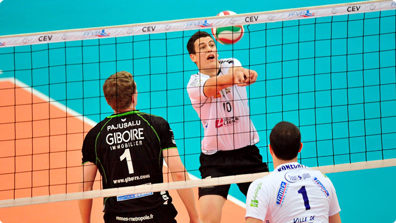 A volleyball player in a black and white uniform jumps to hit the ball over the net, while two opposing team players in black and green, and white and blue uniforms attempt to block. The action takes place on an indoor court.