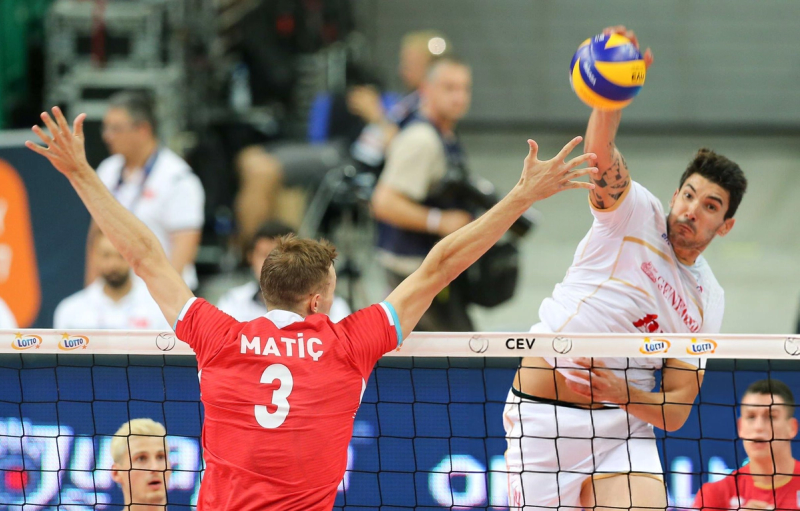 Two volleyball players competing at the net during a match, with one player in white spiking the ball and the other in red attempting a block.