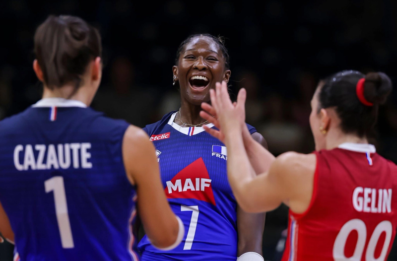 Three female volleyball players celebrating on the court, with one in the center laughing and others clapping hands. They are wearing jerseys with visible names and numbers.